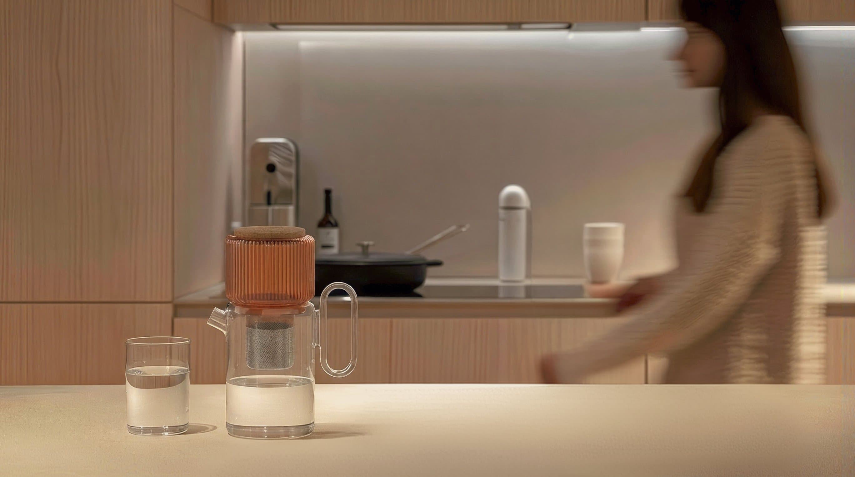 The omomi pitcher and a glass of water on a kitchen counter, someone walking past in the background